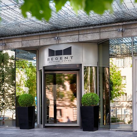 Regent Warsaw Hotel Exterior photo The photo shows the entrance of a building with a modern design. The main feature is a revolving door with a sign above it that reads "REGENT." The entrance is flanked by large potted plants, and the facade consists of glass and reflective surfaces, 