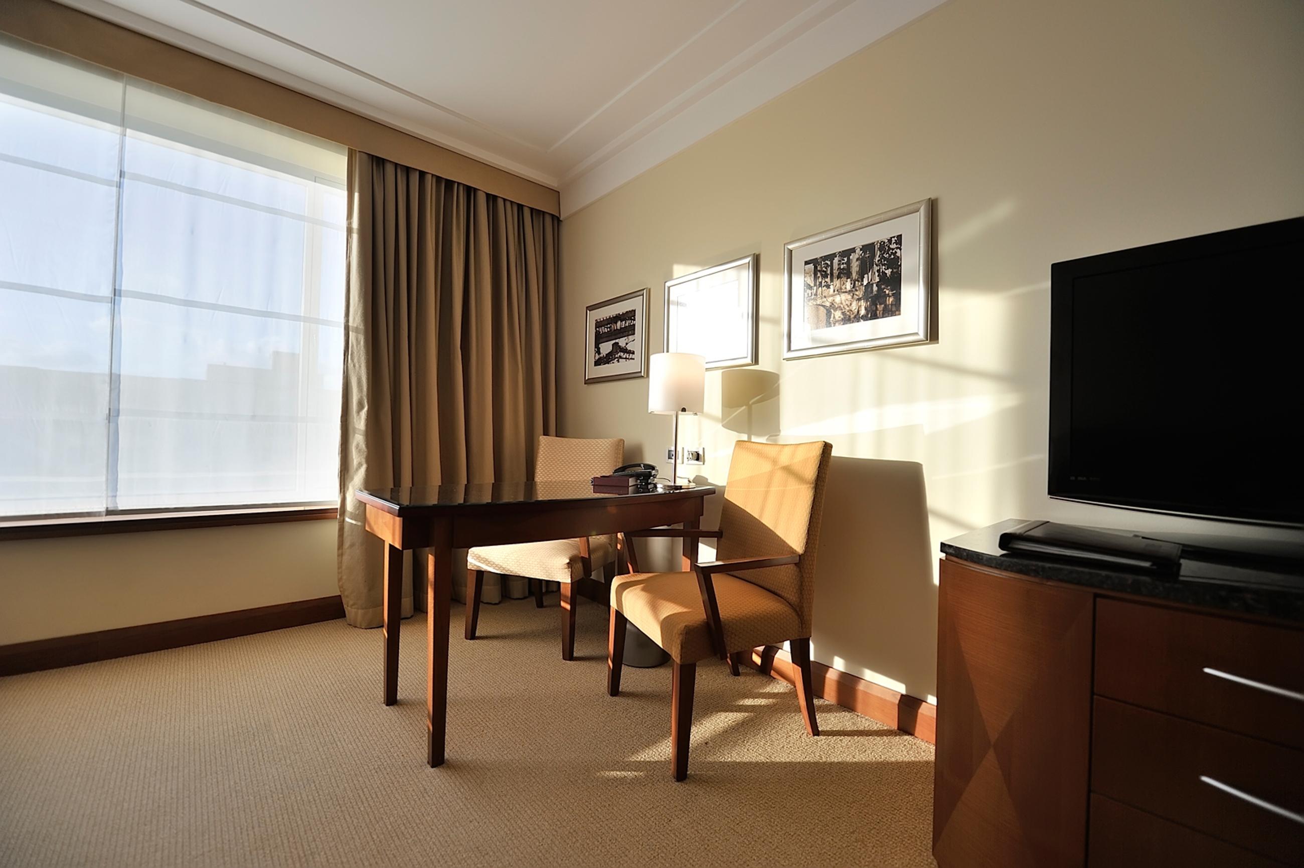 Regent Warsaw Hotel Exterior photo The photo shows a well-lit hotel room or office space. On one side, there is a large window with light-colored curtains that allow natural light to enter. Near the window, there are two upholstered chairs, one of which is positioned at a wooden desk.