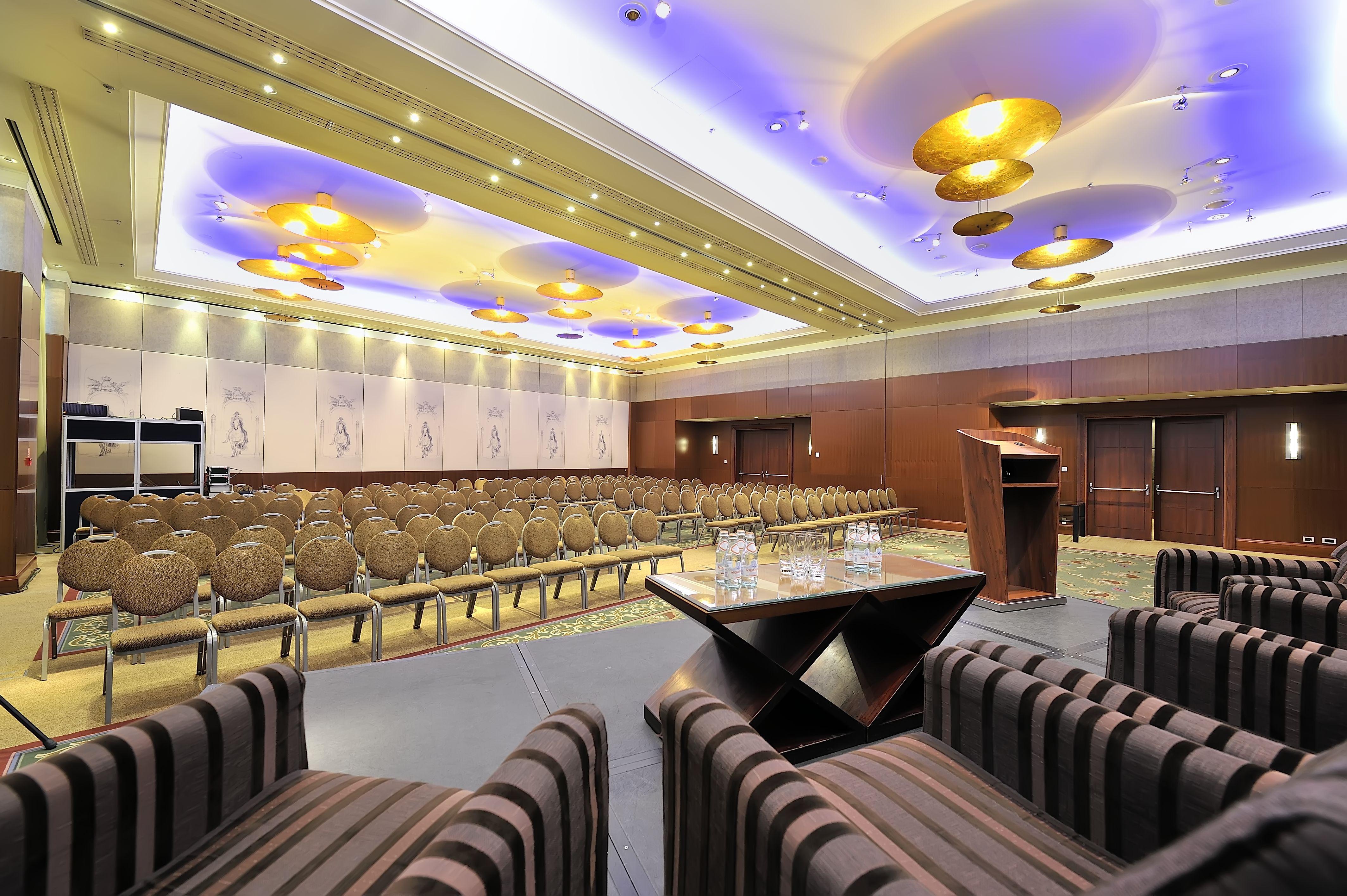 Regent Warsaw Hotel Exterior photo The photo shows a conference room or event space set up for a presentation or meeting. There are several rows of beige chairs arranged in front of a podium, which is positioned on a small stage. The room features elegant lighting, with decorative cei