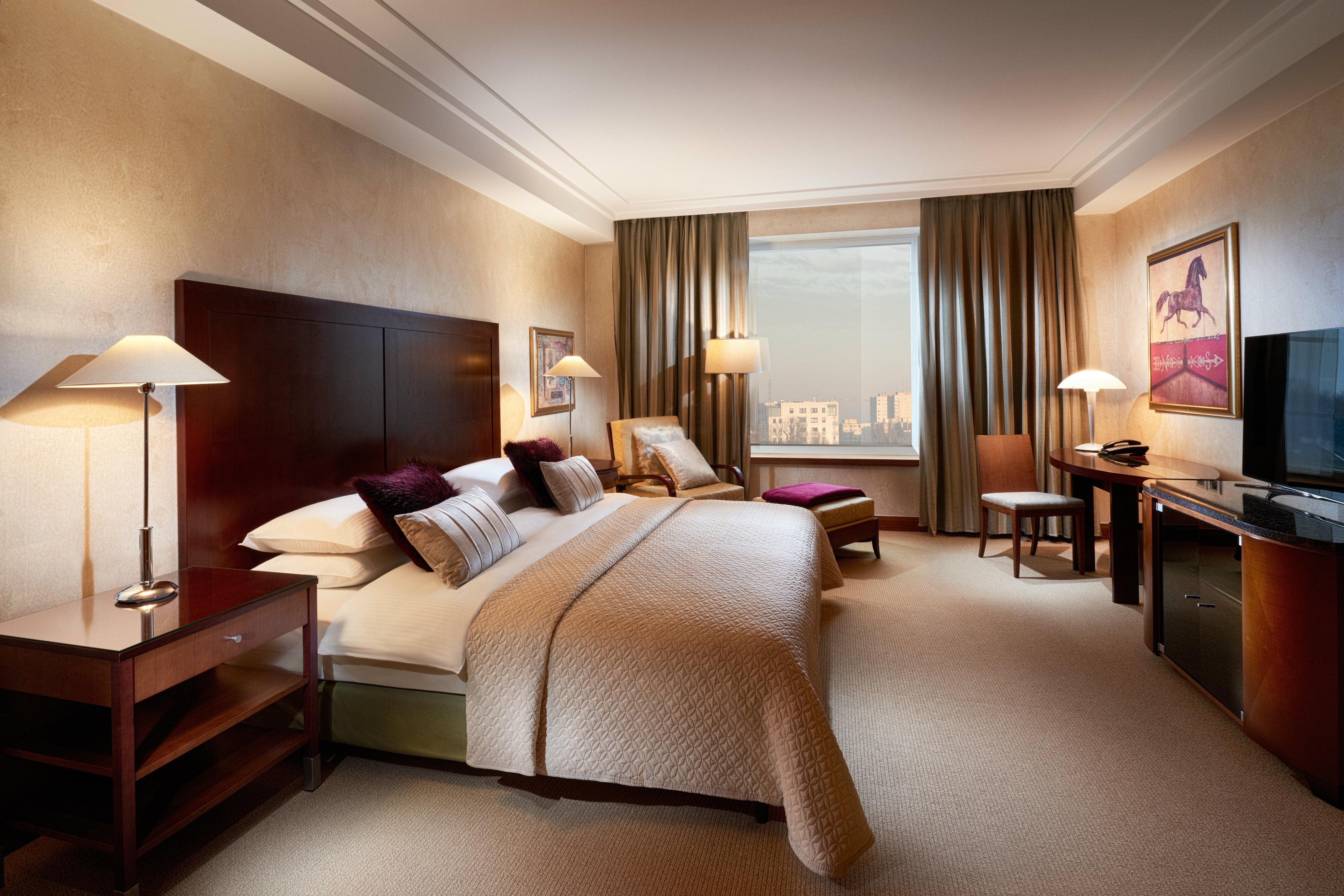 Regent Warsaw Hotel Exterior photo The photo shows a well-appointed hotel room featuring a large bed with white and cream bedding. There are bedside lamps on either side of the bed, and a comfortable armchair is positioned nearby. A window with long curtains allows natural light to en
