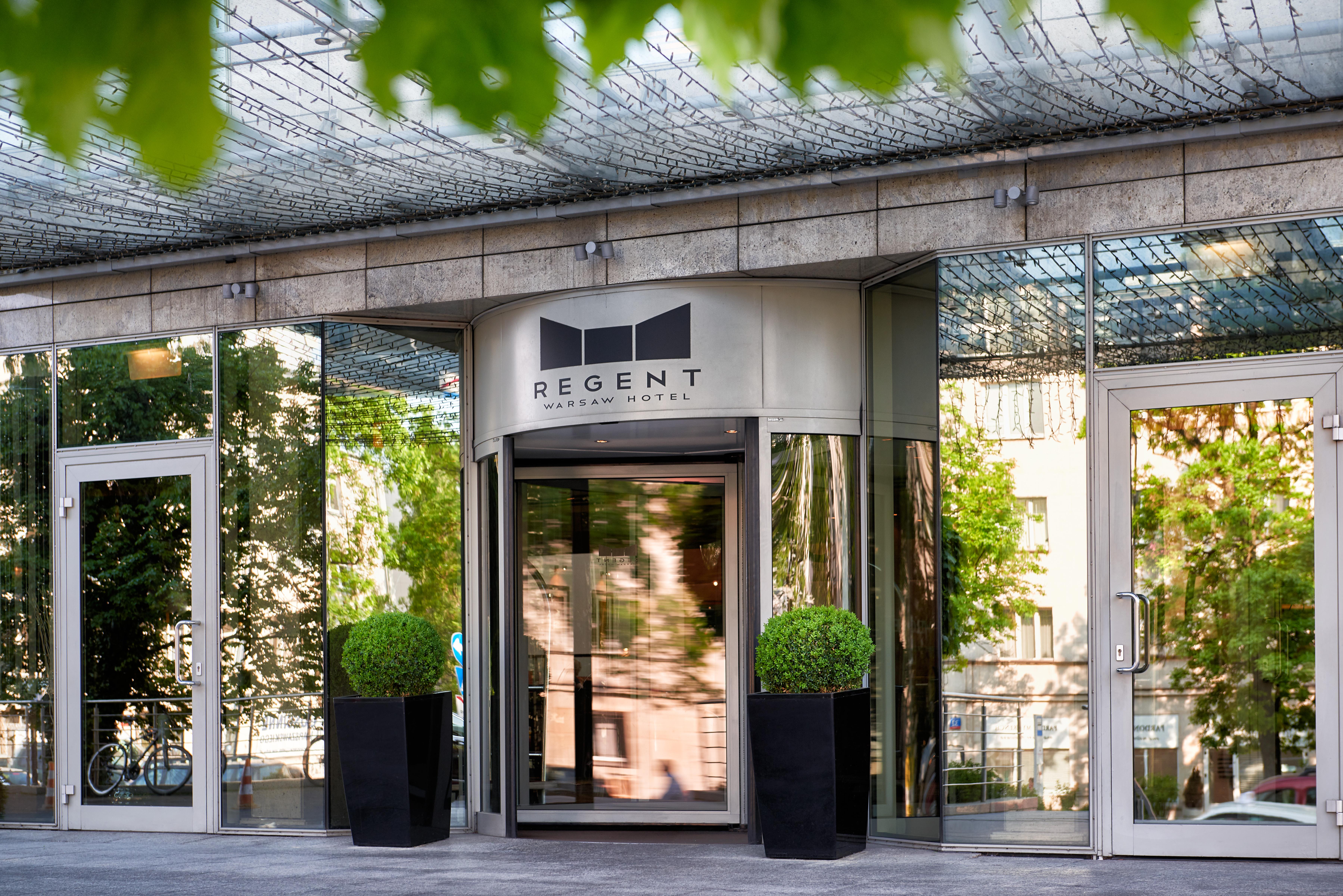 Regent Warsaw Hotel Exterior photo The photo shows the entrance of a building with a modern design. The main feature is a revolving door with a sign above it that reads "REGENT." The entrance is flanked by large potted plants, and the facade consists of glass and reflective surfaces, 