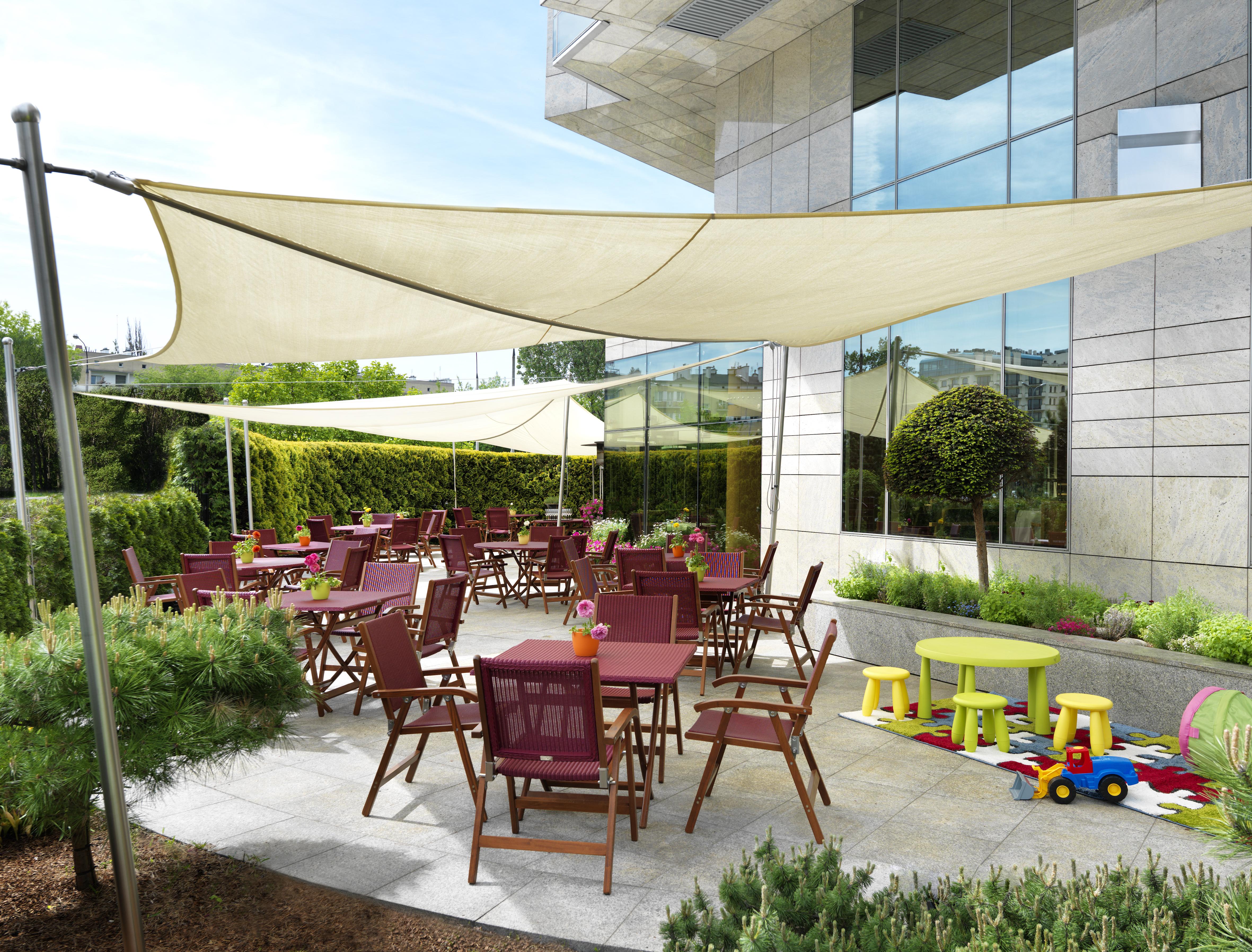 Regent Warsaw Hotel Facilities photo The photo shows an outdoor dining area with a modern design. There are several wooden tables and chairs arranged on a paved surface, with some tables set for dining. Above the seating area, there are large fabric canopies providing shade. In addition