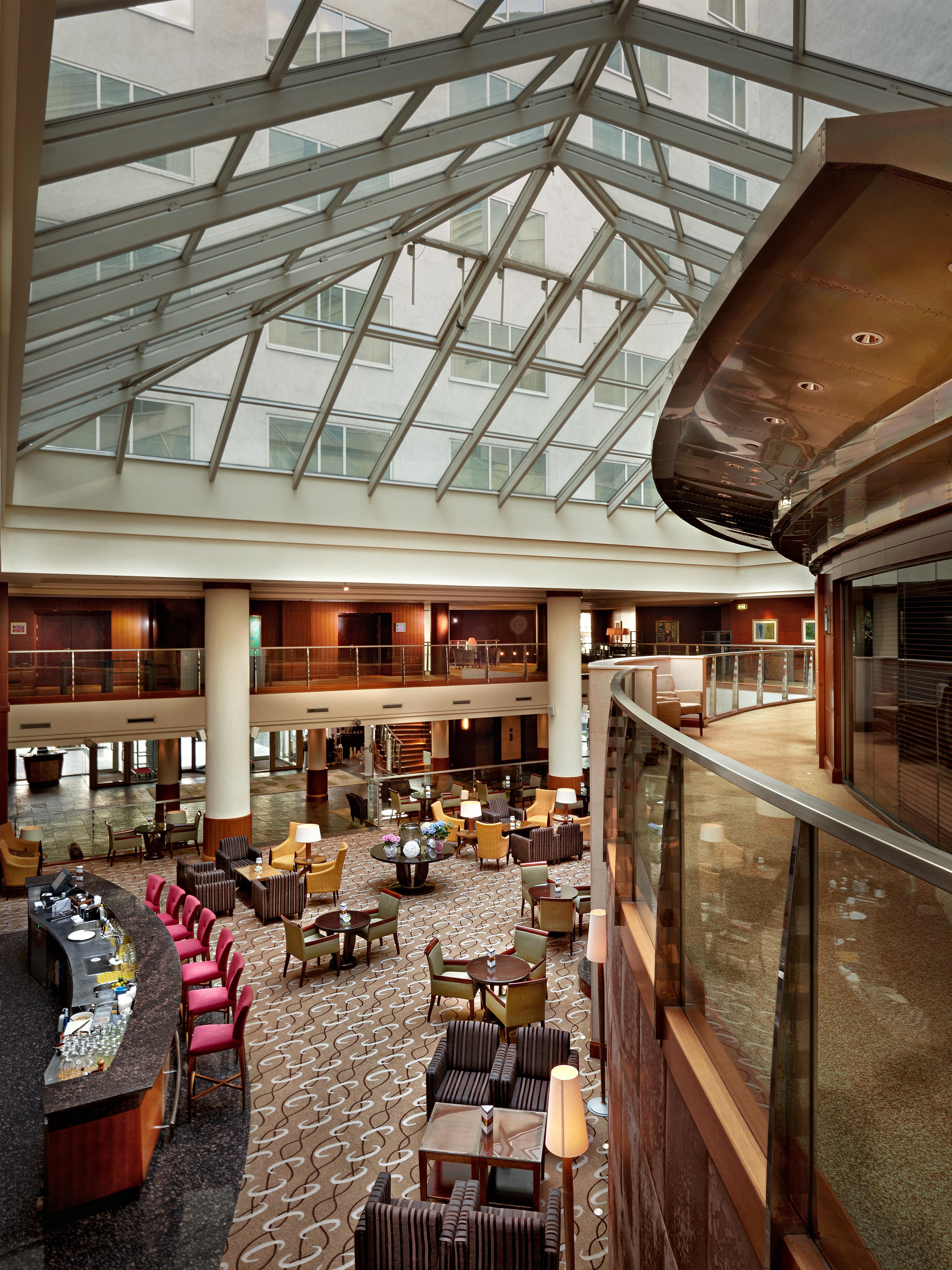 Regent Warsaw Hotel Exterior photo The photo depicts a spacious interior of a hotel lobby or atrium. It features a contemporary design with a high glass ceiling that allows natural light to flood the space. The area is furnished with comfortable seating arrangements, including armchai