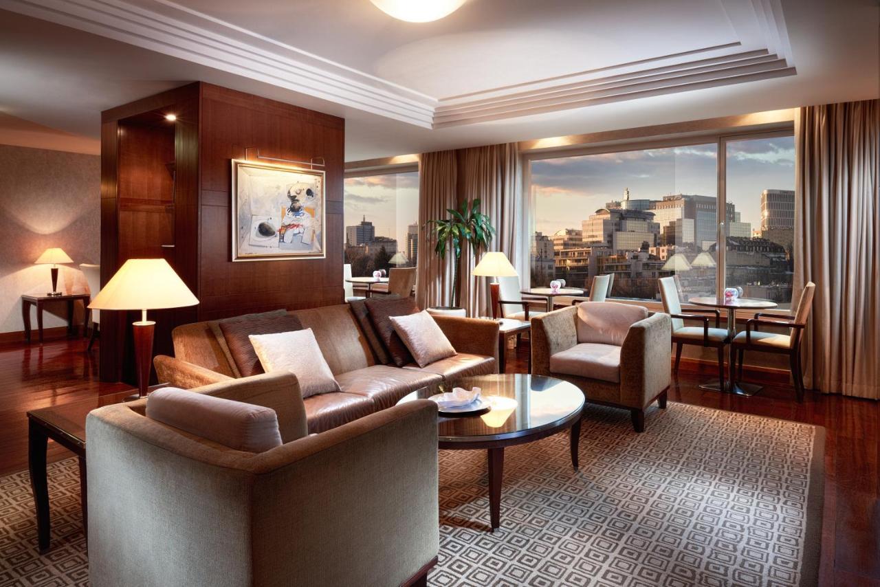 Regent Warsaw Hotel Exterior photo The image shows a stylish and modern living room with large windows offering a view of a city skyline. The room features a comfortable seating area with a sofa, armchairs, and a coffee table. There is a decorative piece of art on one wall, and soft l