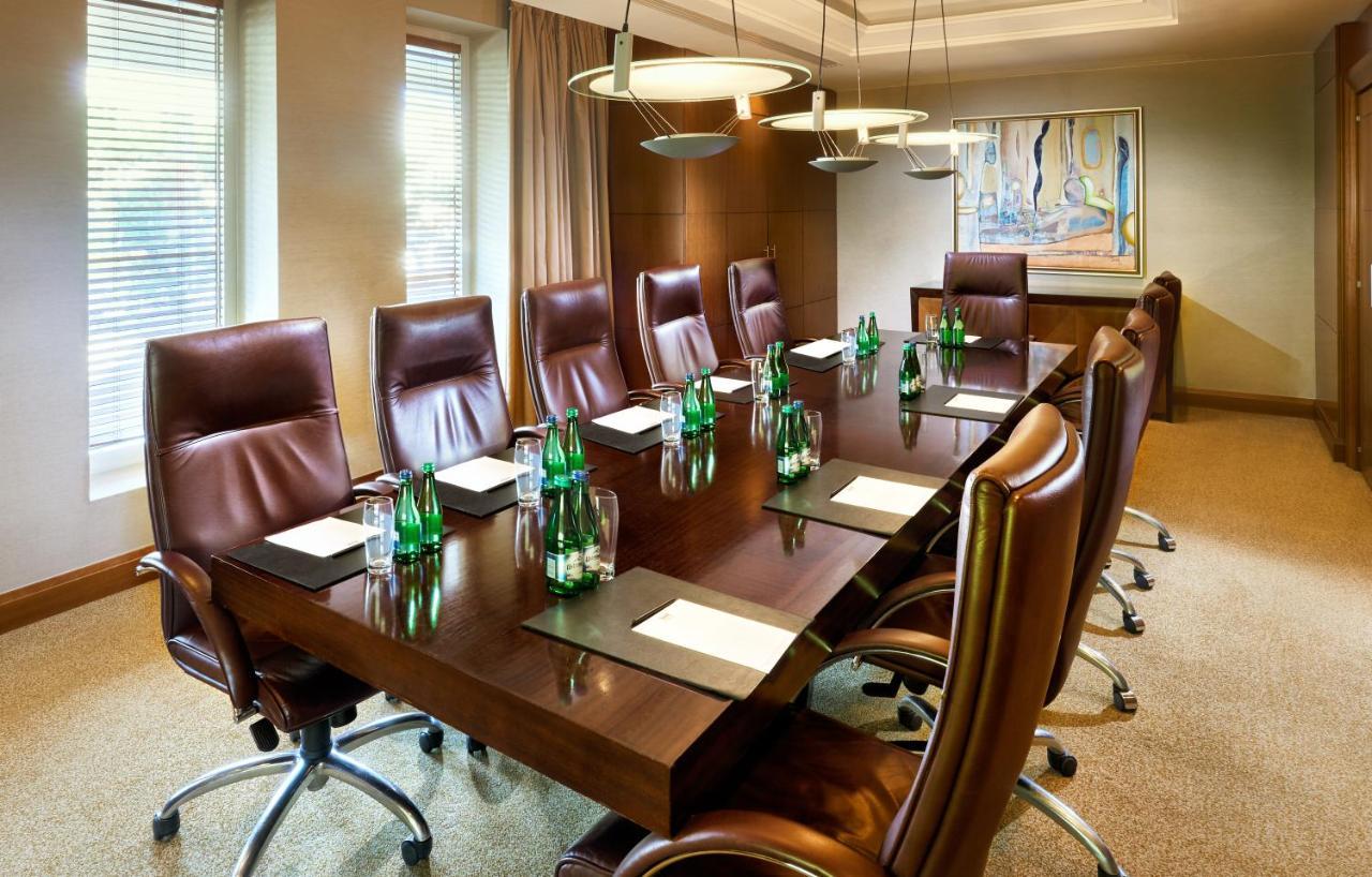 Regent Warsaw Hotel Exterior photo The photo shows a modern conference room with a long wooden table surrounded by several brown leather executive chairs. On the table, there are glass bottles of water and paper notepads, suggesting preparation for a meeting. The room features large w